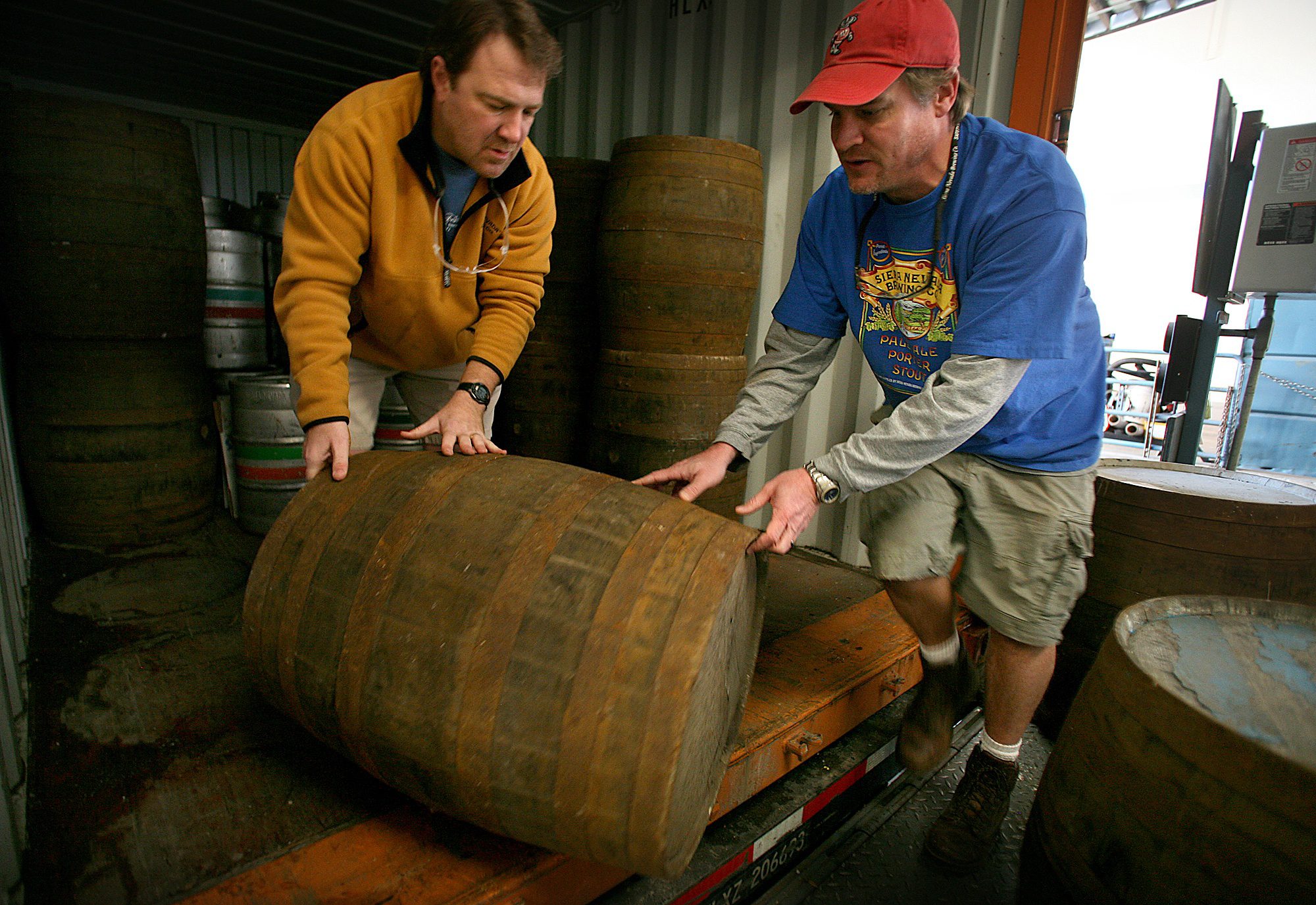 Two male brewers rolling a wooden barrel out of a delivery truck