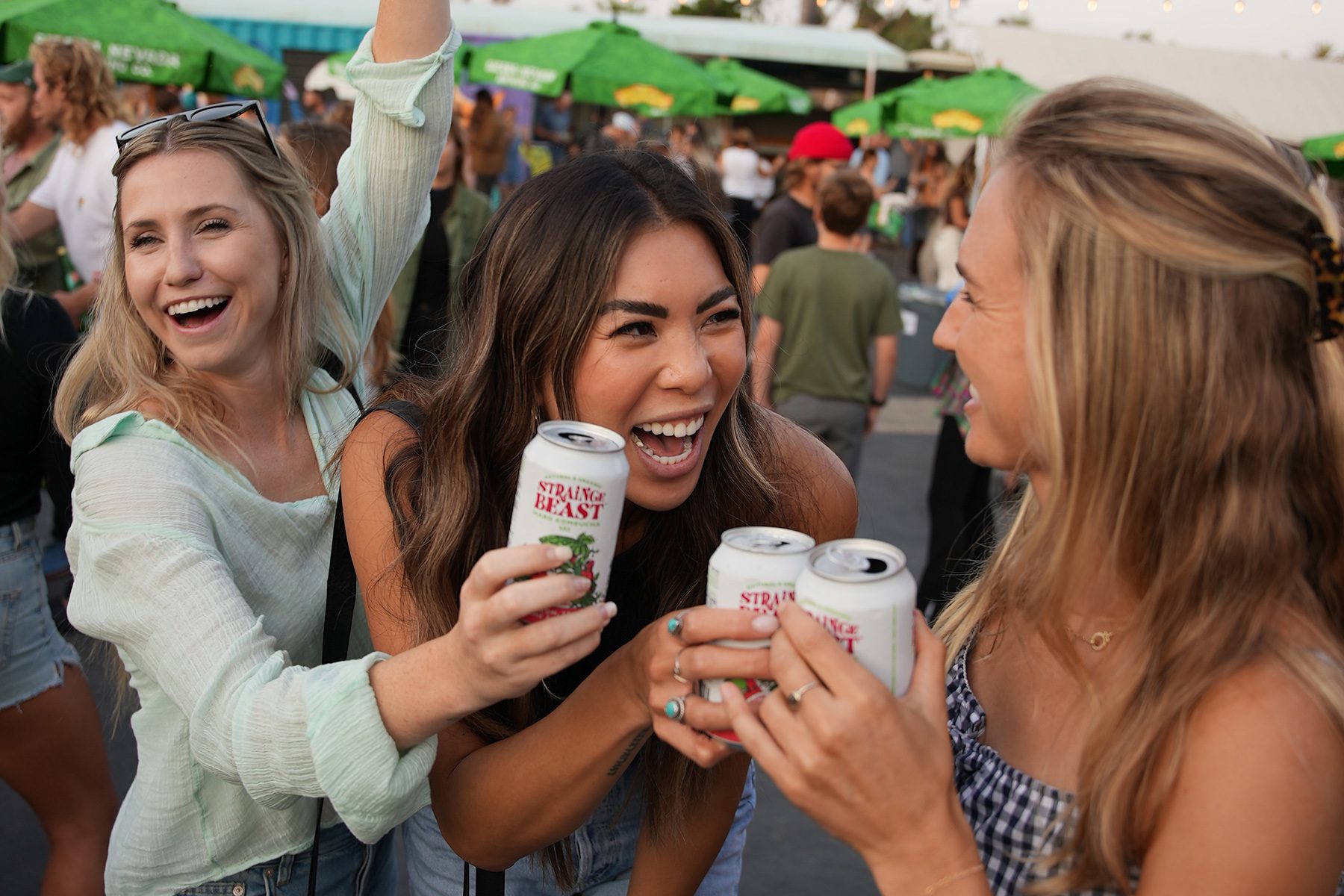 Three friends smiling and toasting cans of Strainge Beast hard kombucha at an outdoor event