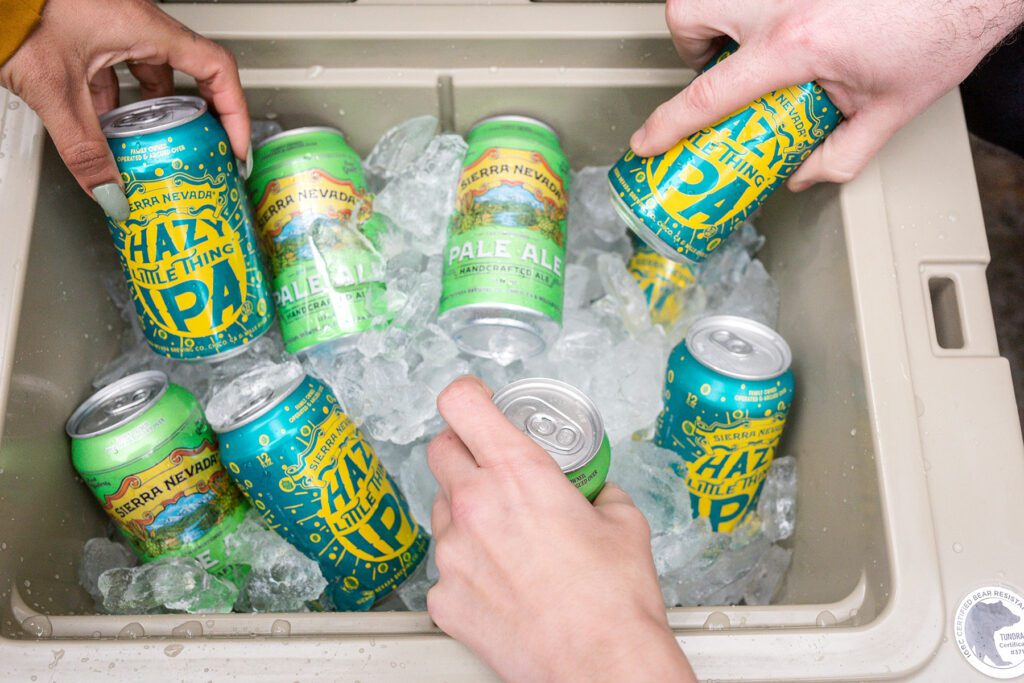 Hands grabbing cans of Hazy Little Thing and Pale Ale from an ice-filled cooler