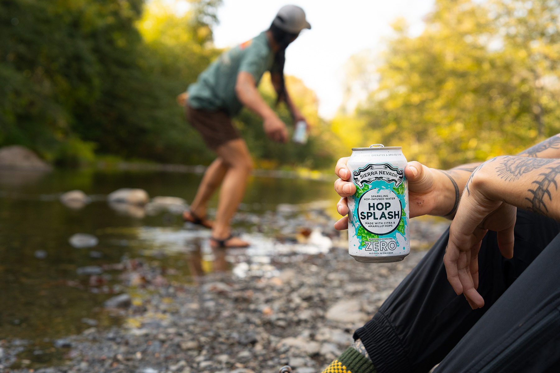 Person holding a can of Sierra Nevada Hop Splash hop water while a friend skips a rock on a creek